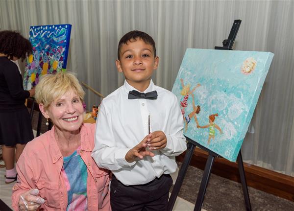 An artist and a student smile at the camera with their picture behind them. Their picture depicts two children swimming on a sunny day.